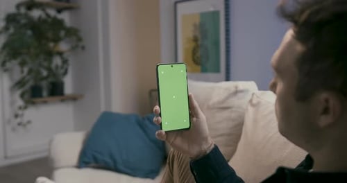 Man holding a smartphone with green screen, looking while sitting in the comfort of his home