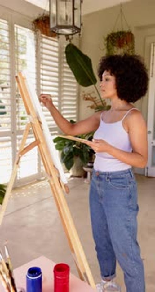 Vidéo verticale : Peinture sur toile, femme debout au chevalet dans une pièce lumineuse avec des plantes
