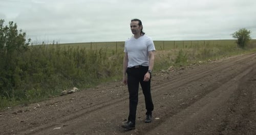 A man dressed in a white t-shirt and dark pants strolls along a dirt road surrounded by expansive gr