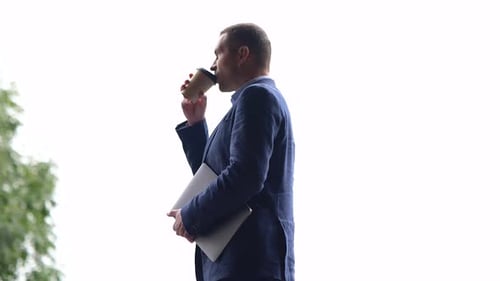 Side View of Caucasian Male Professional Holding Laptop and Drinking Coffee While Standing Outdoors