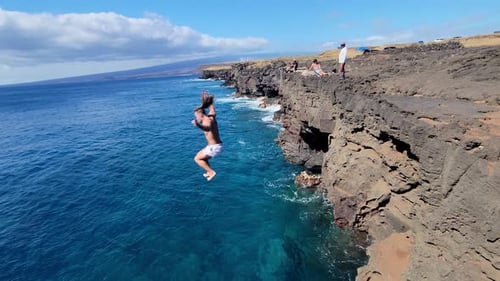 Young man mid-air as he leaps off a rugged seaside cliff into vibrant turquoise water, with onlooker