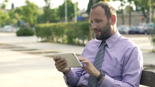 Young Businessman Browsing Internet On Tablet Sitting On Bench In City