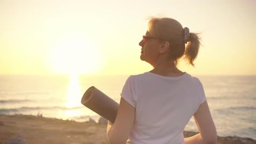 Senior Woman Holding Yoga Mat Enjoying Ocean at Sunset