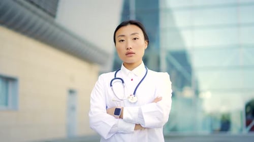 Close up portrait of Asian woman doctor looking at camera with arms crossed on background of modern