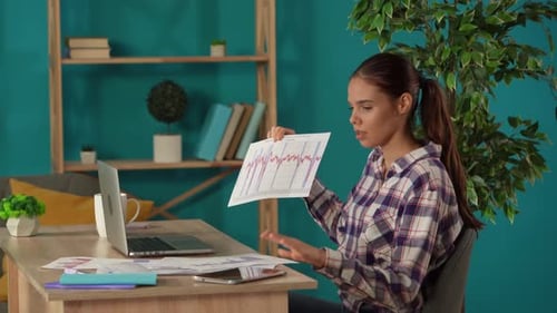 Medium Shot Capturing a Woman Having a Videocall on a Laptop in Her Home Office Discussing Graphs