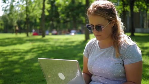 A Young Focused Woman in Sunglasses is Working with a Laptop in a Summer Park