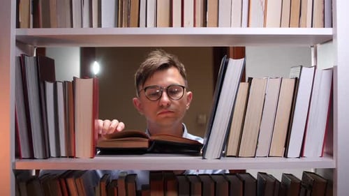 Young Man Reading a Book in a Library Surrounded By Bookshelves