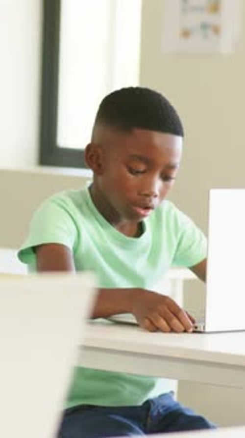 Vertical video: Typing boy glancing up, raising hand for attention at school with laptop, green tee