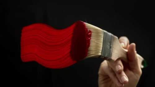 Close-up of a brush with red acrylic paint applied by an artist to glass against a black background.