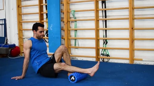 a still shot of a guy massaging his calves using a massage roller inside a gymnastics gym near the