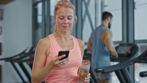 Beautiful Athletic Woman Uses Smartphone Walks Through Fitness Club Gym. Using Social Media, Postin