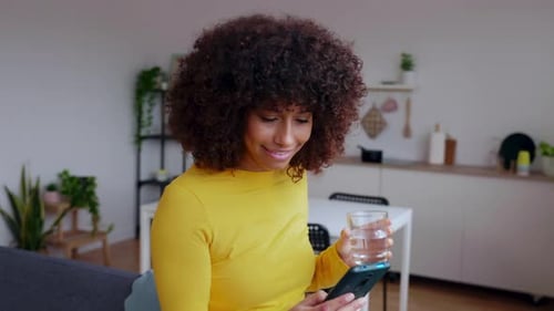 Young Woman Drinking Water While Using Mobile Phone at Modern Loft Apartment