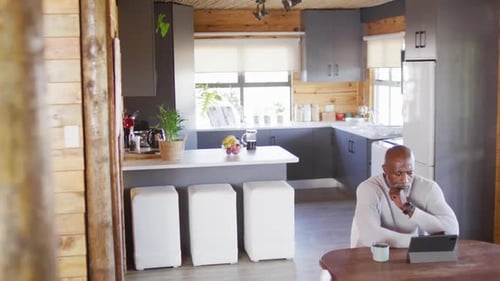 Senior african american man in log cabin, sitting at table and using tablet, slow motion