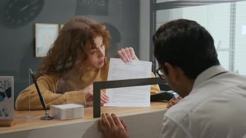 Woman Presenting Document to Male Bank Employee Across Teller Window