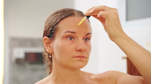 Woman Applying Retinol Serum Drop on Forehead While Looking in Mirror, Front View with Neutral Expre