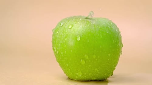 Slow motion shot of water splashing on fresh green apple on beige background.