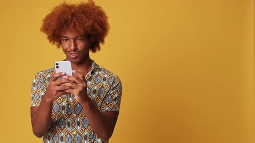 Positive guy uses smartphone, isolated on yellow background in studio