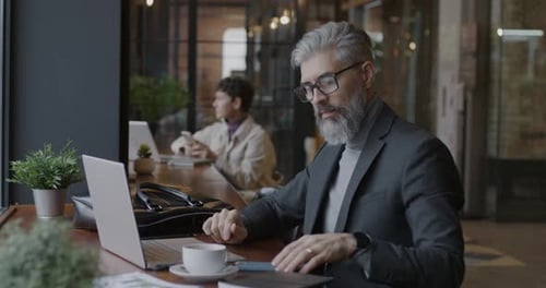 Mature Businessman Working with Laptop Typing Then Speaking on Mobile Phone Discussing Work in Cafe