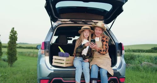 Girl and Woman with Flowers Sit in Car Trunk