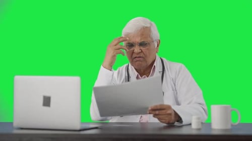 Tensed Indian senior doctor reading health reports Green screen