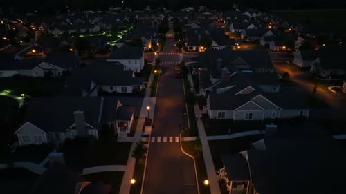 Aerial night view of a quiet suburban neighborhood, illuminated by streetlights and house lights, sh