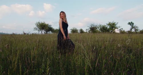 Beautiful female blonde model in black dress stands on a meadow in apple garden