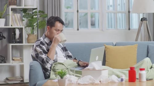 Sick Asian Man With Blanket Drinking Hot Water While Lying On Sofa Works On A Laptop Computer