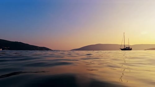 Blick von der ruhigen Meeresoberfläche auf die Silhouette der Yacht vor Sonnenuntergang, Himmel und Bergen