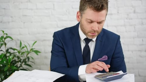 Ein gestresster Geschäftsmann, der einen Taschenrechner verwendet, um Schulden und Steuern mit Stapeln von Finanzkonten zu berechnen