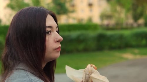 Profile Footage of Caucasian Woman with Long Dark Hair Chewing Cheeseburger Resting in the Park