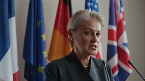 Woman Speaking at Press Conference with Flags Behind Her