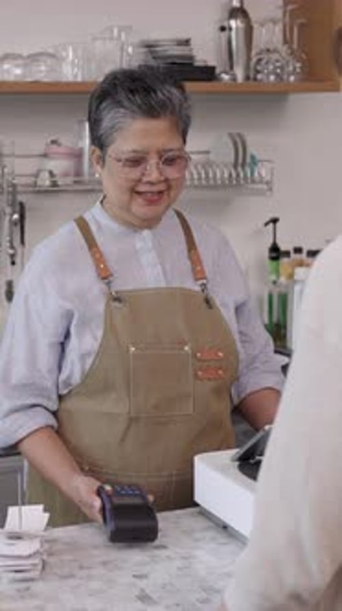 Asian senior female cafe owner wearing apron smiling while receiving payment from customer at coffee