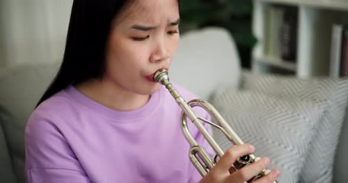 A young woman practices playing the trumpet while sitting on a sofa in a cozy living room.