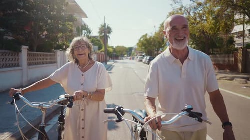 Senior Couple Walks Bikes Down Suburban Street Smiling