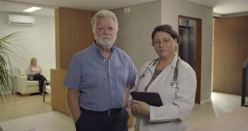 Senior man and female doctor standing together in clinic hallway, facing camera with calm and