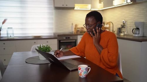 Businesswoman multitasking at her home office, talking on a smartphone and writing in a notebook