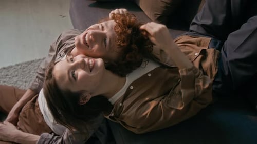 Joven sonriente acostada en el sofá y abrazando a su novio, sentada en el suelo