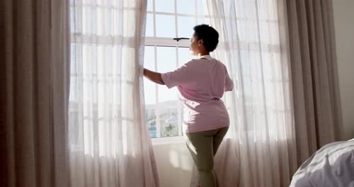 African American woman opens curtains to a sunlit room at home