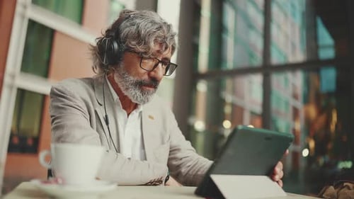 Mature businessman and headphones, sits in an outdoor cafe and works using tablet