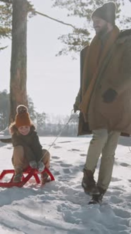 Cheerful Young Caucasian Father Pulling Daughter on Sled in Snowy Forest