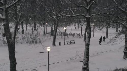 Streetlamps illuminate snow falling on people crisscrossing through an urban winter park, aerial pus
