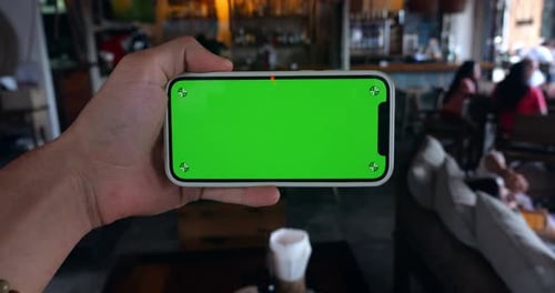 Man's Hand Holding Smartphone in Horizontal Position with Green Screen Chroma Key Indoors at Cafe or