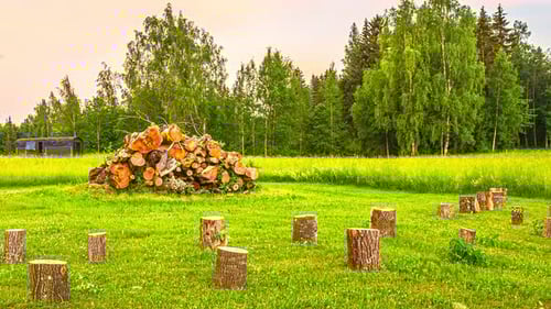 People Place Wooden Table And Chair On The Grass With Tree Logs. Pile Of Woods Burn At Sunset. time