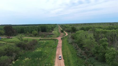 Following aerial fly over a car driving along a rural dirt country road through green meadows in