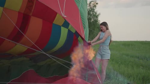 Woman Grips Hot Air Balloon Envelope Near Burning Burner Flame in Green Field
