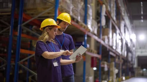 Two warehouse workers, wearing protective helmets and identification badges, collaborate, consulting