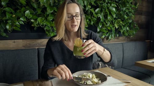 Woman Drinking Juice And Eating Salad Sitting On Sofa In Cafe