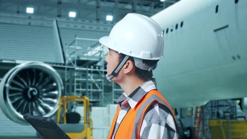 Side View Of Asian Male Engineer Looks At Tablet, Looks Around While Aircraft Maintenance