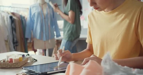 Zwei asiatische Modedesignerinnen mit einem digitalen Tablet für den Gelegenheitsgebrauch kreieren in ihrem Geschäft eine neue Sammlung.