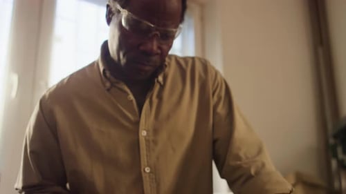 Black Woodworker Cutting Plank with Handsaw at Workbench